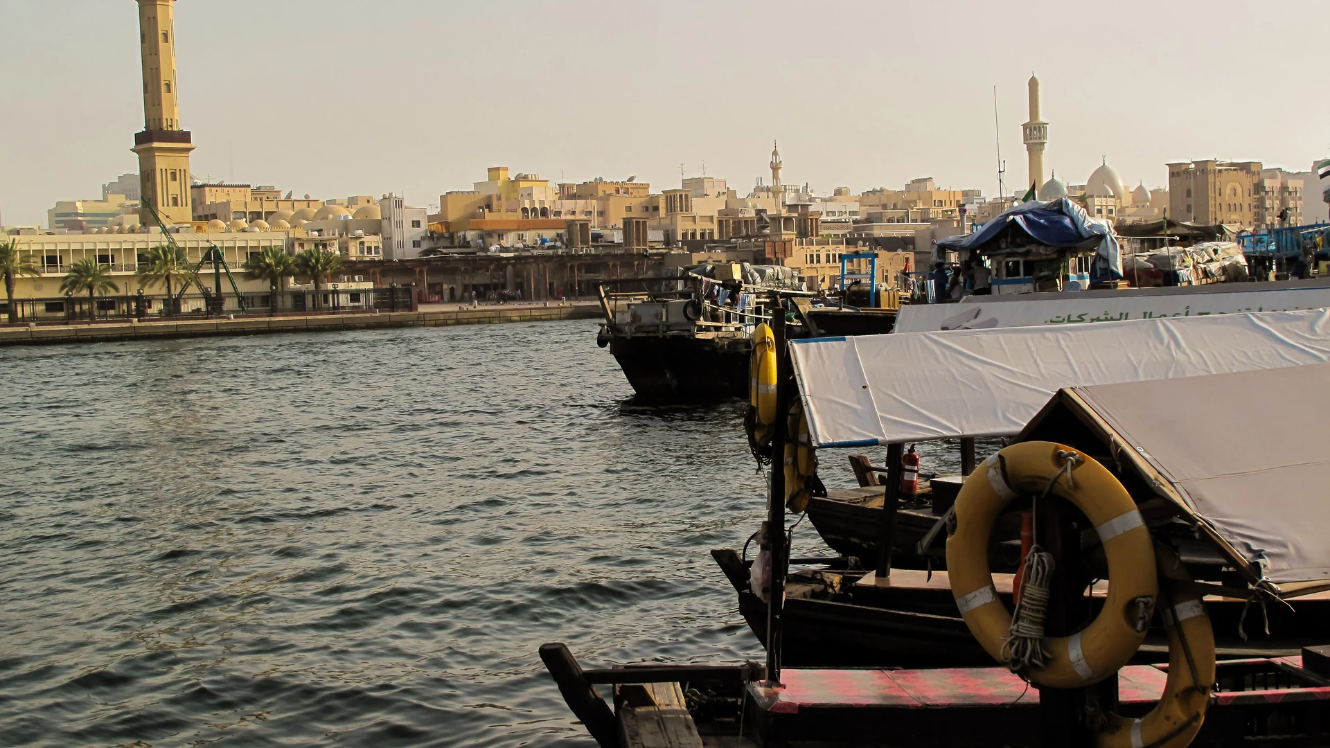 Wooden boats moored on Dubai Creek in the late-afternoon haze, with a minaret and sand-coloured buildings of Old Dubai across the water