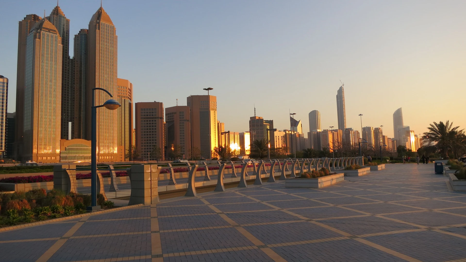The paved Corniche promenade in Abu Dhabi at sunset with the golden towers of the downtown skyline behind it