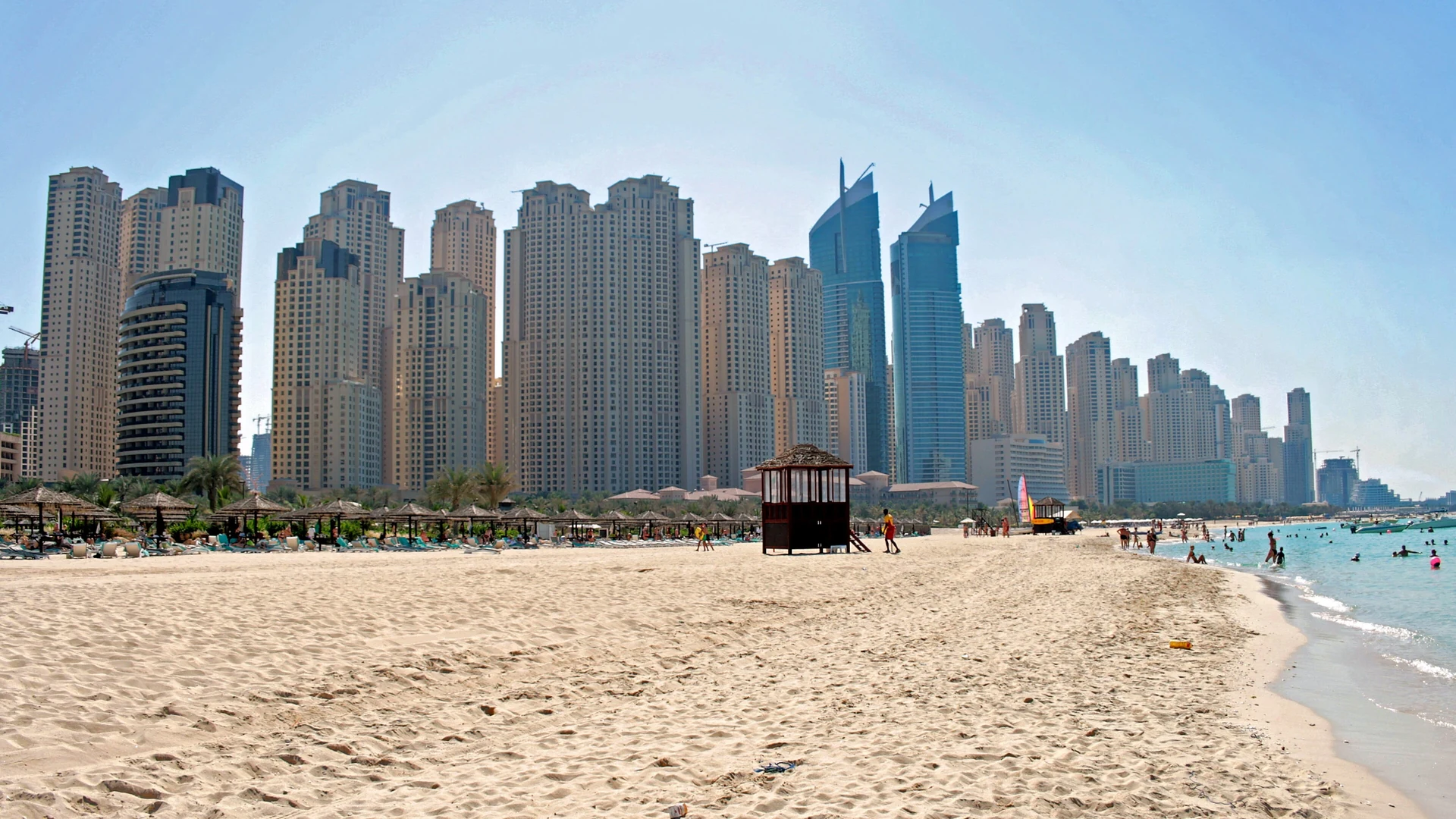 A wide sandy beach at Jumeirah Beach Residence in Dubai under a clear sky, with the JBR apartment towers behind and swimmers in the turquoise sea