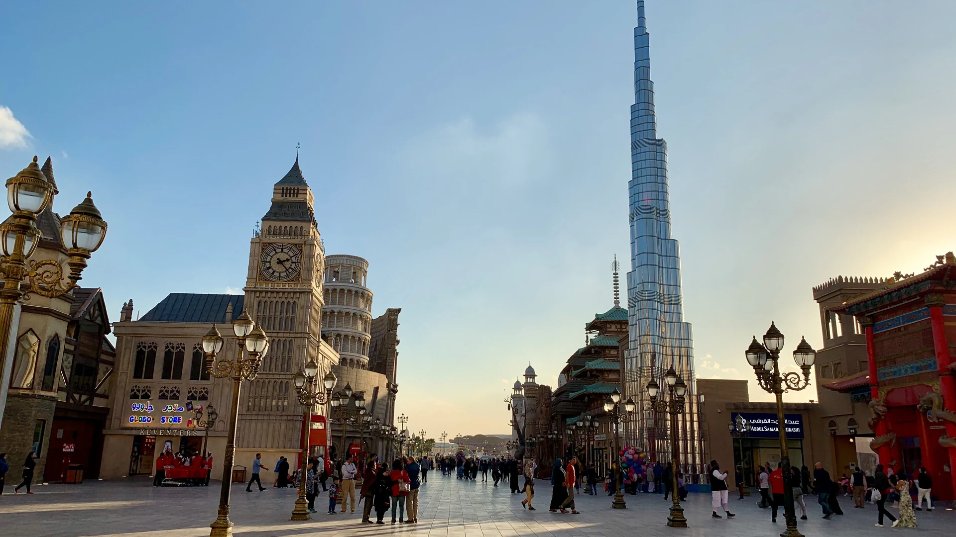 Visitors on a Global Village walkway in Dubai at golden hour, with a Big Ben replica and the Leaning Tower of Pisa beside a Chinese-style pavilion