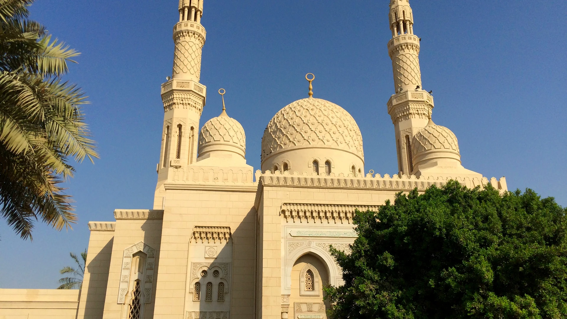 The cream-coloured domes and two tall minarets of Jumeirah Mosque in Dubai against a clear blue sky, with palm fronds in the foreground